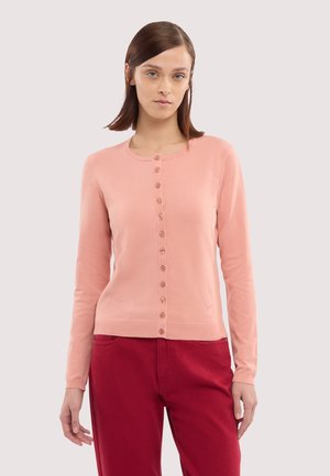 Donna che indossa un cardigan rosa chiaro abbottonato e pantaloni rossi, in piedi contro uno sfondo chiaro uniforme, rivolta verso l'obiettivo.