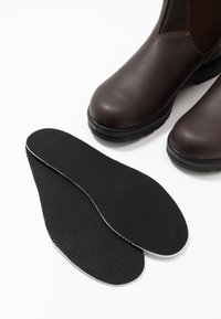 Botas de cuero marrón hasta el tobillo con una plantilla negra texturizada colocada junto a ellas, que presenta un borde blanco en el borde.