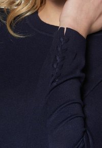 Manche de pull côtelé bleu marine avec détail tressé et petite encoche près du poignet, portée par une personne aux cheveux blonds.