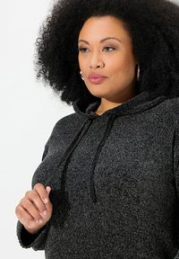 Donkergrijze hoodie met een getextureerd ontwerp, voorzien van een trekkoord bij de hals en een relaxte pasvorm. De hand van het model rust op de stof.
