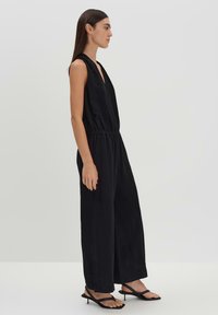 Black sleeveless jumpsuit with a deep V-neck, elastic waist, wide legs, and a smooth texture. Worn with black strappy sandals.