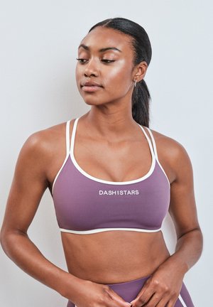 Donna con capelli scuri raccolti in una coda di cavallo che indossa un reggiseno sportivo viola con rifiniture bianche e il testo "DASH AND STARS,", guardando leggermente a sinistra.