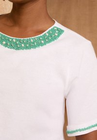 Blouse blanche à manches courtes mettant en valeur un col en perles turquoise avec des motifs intrigants de perles petites et moyennes, offrant un détail texturé.