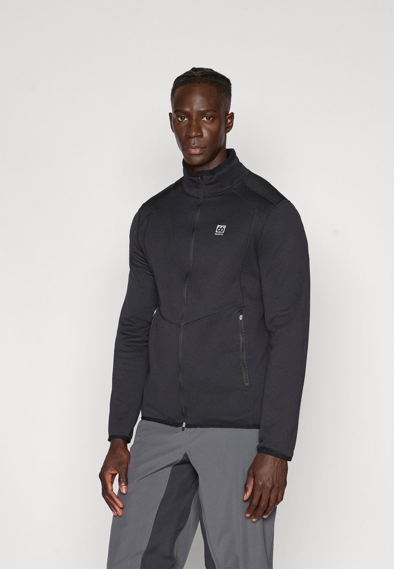 Zwarte zip-up jack met hoge kraag, gemaakt van zachte stof. Voorzien van twee met rits afgesloten zakken en een logopatch op de linkerkant van de borst.