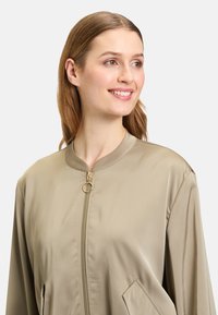 Giacca bomber in satin beige chiaro con collo rotondo, zip dorata e due tasche laterali per un aspetto pulito. Nessun motivo visibile.