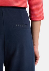 Námornícke modré nohavice vyrobené z hladkej tkaniny, s vreckom na zadnej strane a logom "ELBSAND". Čiastočne viditeľný pruhovaný červený top.