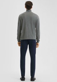 Grijze lange mouwen coltrui met een gestructureerde afwerking, gecombineerd met marineblauwe broek. Model staat met de rug naar de camera, met zwarte schoenen aan.