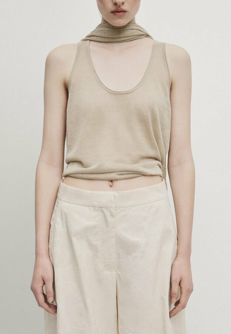 Femme portant un haut beige sans manches et un pantalon beige clair taille haute, debout devant un fond uni.