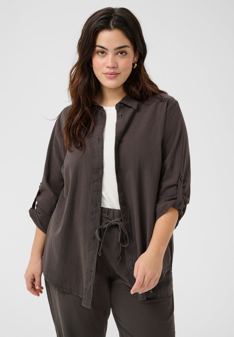 Chemise boutonnée en charbon foncé avec un tissu doux et texturé ; elle présente des manches roulées et une coupe décontractée. Associée à un pantalon à cordon de serrage assorti.