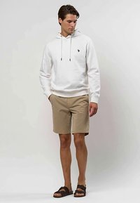 Homme portant un sweat à capuche blanc, un short beige et des sandales noires, debout devant un fond clair uni, les mains dans les poches.