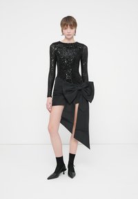 Crni tijelac dugih rukava sa šljokicama uparen s crnom suknjom koja ima veliki mašnu u struku. Uski kroj, elegantan dizajn, uz cipele s oštrim vrhom.