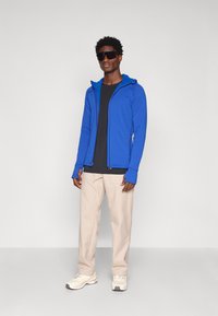 Sudadera azul con cierre, capucha, mangas largas y agujeros para los pulgares, llevada sobre una camiseta negra. Pantalones beige y zapatillas blancas completan el conjunto.
