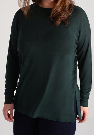 Femme portant un haut à manches longues vert foncé avec des fentes sur les côtés et un pantalon foncé, debout devant un fond clair.