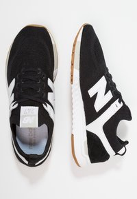 Sneaker New Balance 247 in bianco e nero con tomaia in rete, logo bianco e suola in gomma, mostrati da viste dall'alto e di lato su sfondo bianco.
