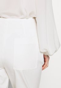 Personne portant un pantalon blanc taille haute avec une poche arrière et un chemisier blanc à manches longues bouffantes.
