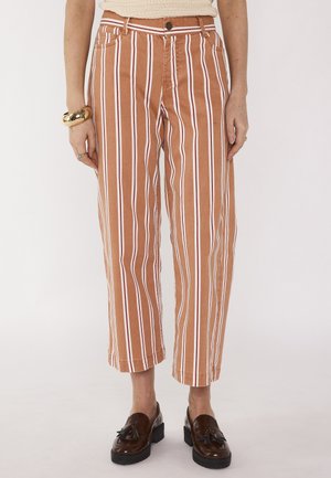Donna che indossa pantaloni corti a vita alta con righe verticali marroni e bianche, mocassini marroni con nappine, una maglietta beige e braccialetti dorati.