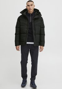 Sort pufferjakke med høj krave, lynlås og elastiske manchetter. Bæres over en mørk top og mørke bukser, sat sammen med blå sneakers.