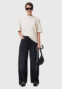 T-shirt crema de corte amplio con un logo sutil, combinado con jeans negros de cintura alta que cuentan con tachuelas laterales y un ajuste relajado, accesorios negros.