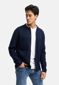 Marineblauer Cardigan aus weichem Stoff, mit einem steifen Kragen, Knopfverschluss und zwei vorderen Taschen, über einem weißen Shirt getragen.