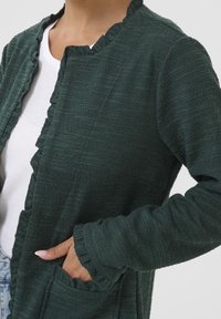 Zelený strukturovaný kardigan s volánovým límcem a manžetami, vybavený kapsami a volným střihem. Látka má tkaný, bohatý vzhled.
