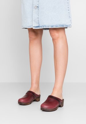 Femme portant des sabots en cuir bordeaux avec des talons en bois et une jupe en denim bleu clair, se tenant sur un sol gris uni.