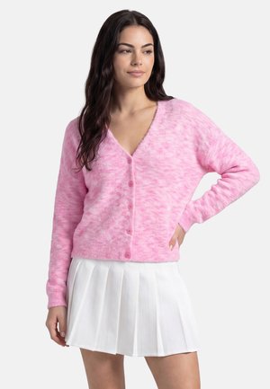 Donna con lunghi capelli scuri che indossa un cardigan rosa peloso con bottoni e una gonna plissettata bianca, in piedi con una mano sul fianco.