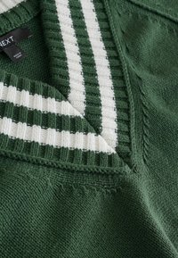 Vihreä neulepaita, jossa on V-pääntie, valkoiset ribbikuvioidut raidat kauluksessa, sekä teksturoitu kangas teksturoidulla pinnalla.