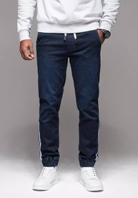 Joggers azul oscuro con puños elásticos, rayas blancas a los lados y cintura con cordón. Combinados con zapatillas blancas para un look casual.