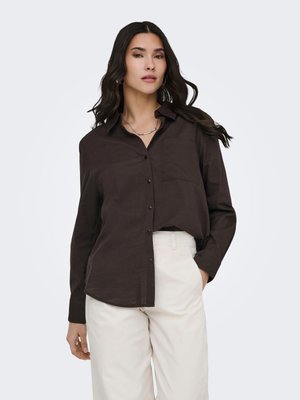 Femme aux longs cheveux foncés portant une chemise marron foncé à boutons et un pantalon beige clair, debout, une main dans la poche.