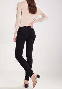 Jeans neri slim-fit con dettagli sulle tasche, abbinati a un top beige chiaro e testurizzato con maniche arricciate. Indossati con tacchi beige.