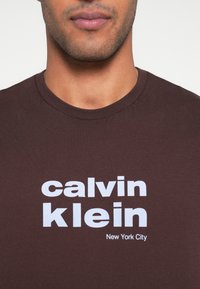 Tricou maro din bumbac cu text alb îndrăzneț: „calvin klein” și „New York City” într-un font modern. Guler rotund și mâneci scurte.