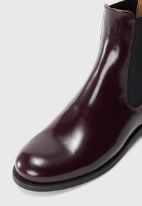 Bottine Chelsea en cuir bordeaux brillant avec un panneau latéral élastique noir et une semelle noire sur un fond clair.