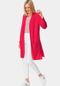 Cardigan long rouge en tissu doux avec un devant ouvert et des poches, associé à un haut rayé et un pantalon blanc. Modèle portant des baskets blanches.