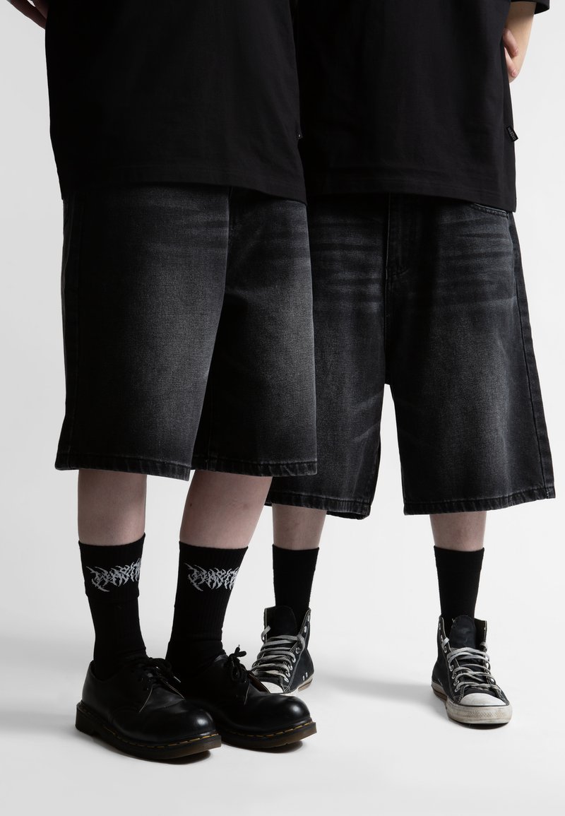 Δύο άτομα που φορούν μαύρα φαρδιά σορτς, μαύρες κάλτσες με λευκά γραφικά, μαύρα παπούτσια και μαύρα τοπ, στέκονται δίπλα-δίπλα.