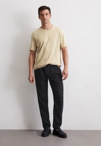 Beige T-Shirt mit kurzen Ärmeln kombiniert mit locker sitzenden schwarzen Hosen und dunklen Slippern. Glatte Textur, einfaches Design und lässiger Schnitt.