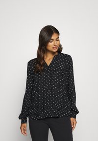 Musternäit must blouse valgete täppidega, pikkade varrukatega, V-kaelusega ja nööbikaunistustega, valmistatud kerge materjali ja pehme tekstuuriga.