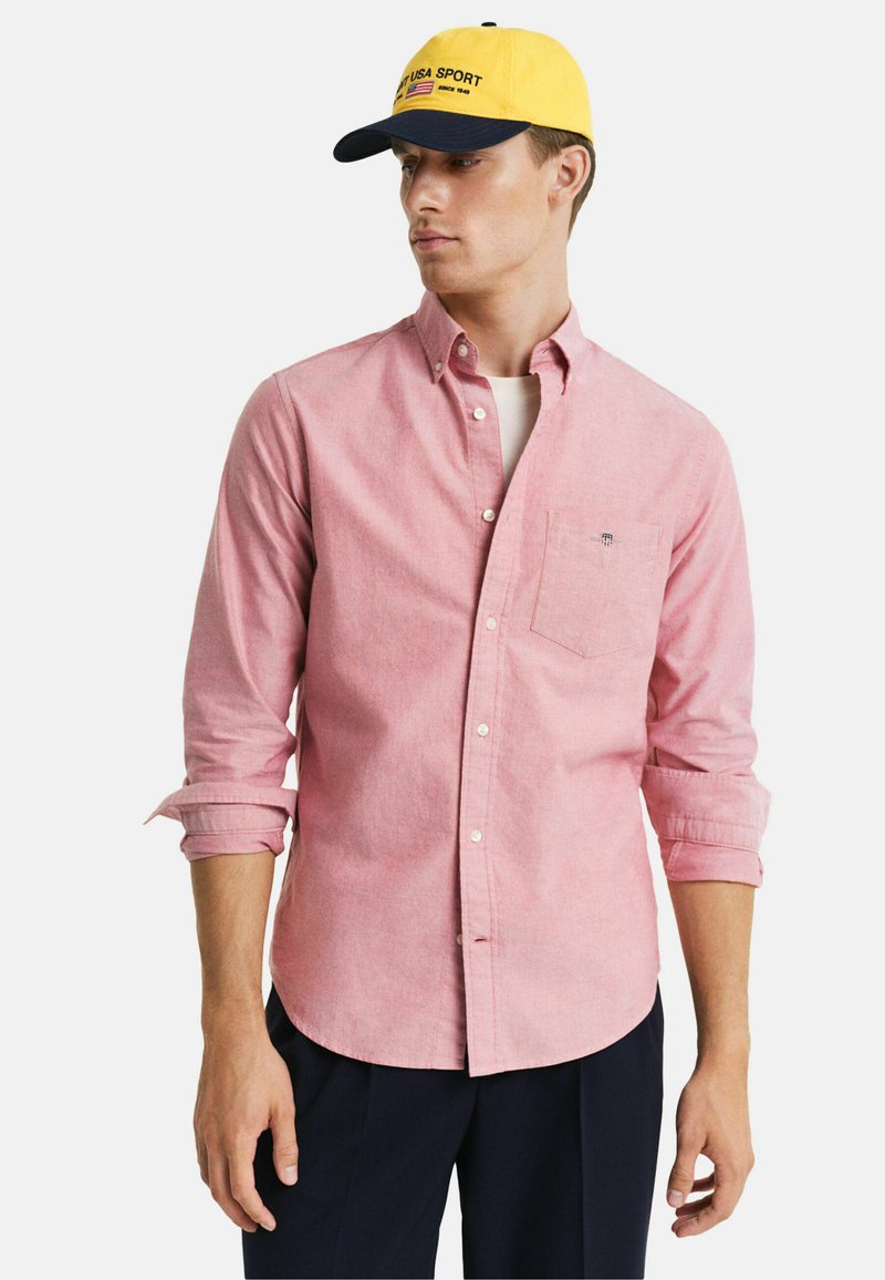 Camicia button-up rosa chiaro con maniche lunghe e taschino sul petto, abbinata a un cappellino giallo con visiera blu navy e scritte.