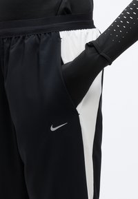 Άτομο που φοράει μαύρο και λευκό παντελόνι Nike, με το χέρι στην τσέπη και μαύρο μακρυμάνικο τοπ με λευκό πουά στο μανίκι.