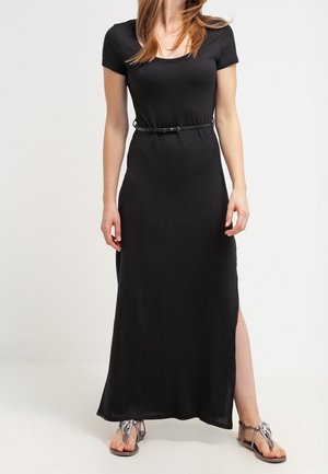 Schwarzes Maxikleid mit kurzen Ärmeln, rundem Ausschnitt und seitlichem Schlitz, in der Taille mit einem dünnen schwarzen Gürtel betont. Dazu flache Sandalen getragen.