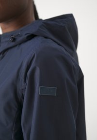 Marinblå lättviktsjacka med texturerad tyg, dragsko i huva och en CMP-logotyp på överarmen.