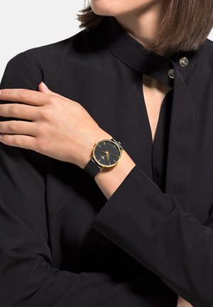 Black watch with a round face, gold accents, and black rubber strap, worn on a hand against a black top with a unique collar design.