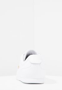 Bílá teniska s hladkou texturou, zaobleným tvarem a polstrovaným límcem. U paty malá zlatá značka. Pryžová podrážka.