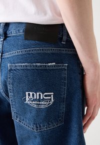 Jean en denim bleu foncé avec une coupe droite, arborant une étiquette en cuir à la taille et un logo brodé blanc sur la poche arrière.