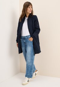Giacca trapuntata blu marina con tasca zip, abbinata a jeans azzurri e scarpe bianche con logo nero. Tessuto liscio, design fino al ginocchio.