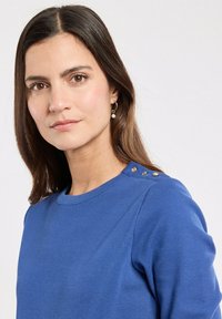 Blauwe gebreide trui met een ronde hals en schouderknopen. Heeft een geribbelde textuur en een aansluitende pasvorm.