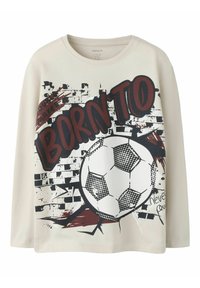 Kremfarget langermet skjorte med en stor grafikk av en fotball mot et slitt murveggdesign med røde og svarte aksenter.