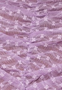 Tissu en dentelle de lavande avec de petits motifs floraux en relief et des détails de points zigzag, présenté en doux plis et en gros plan sur la texture.