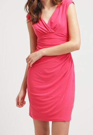 Vrouw die een nauwsluitende, mouwloze roze jurk draagt met een V-hals en gedetailleerde taille, staand tegen een effen witte achtergrond.