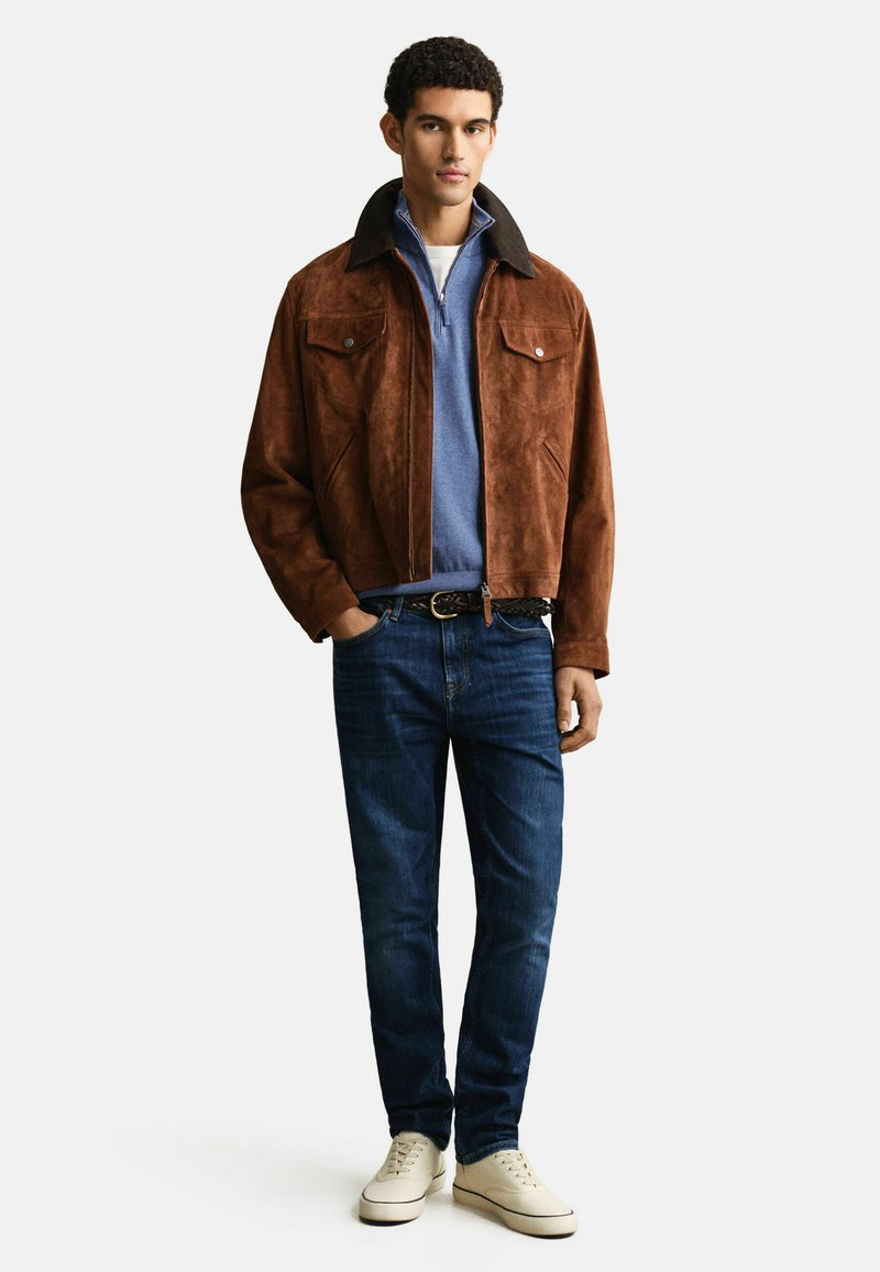 Mann trägt eine braune Wildlederjacke über einem blauen Pullover mit Viertelreißverschluss, dunkelblaue Jeans, geflochtener Gürtel und weiße Turnschuhe, steht vor weißem Hintergrund.