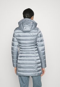 Mujer vestida con una chaqueta acolchada ligera de color azul con capucha, de pie de espaldas contra un fondo liso.